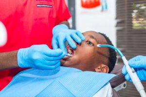 Dentist in red scrubs and blue gloves examines a patient's teeth with dental tools in a clinic setting