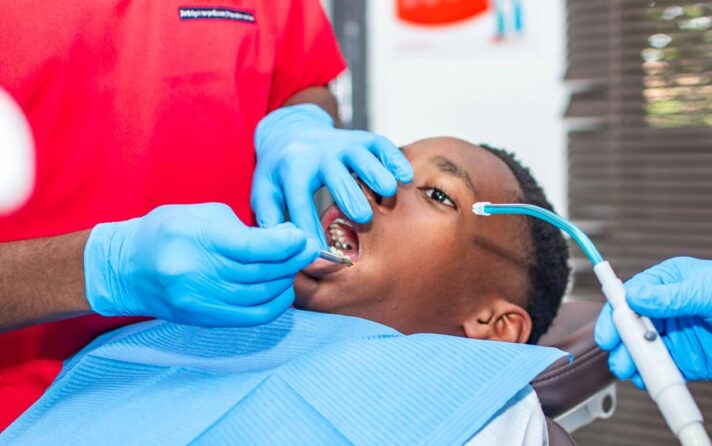 Dentist in red scrubs and blue gloves examines a patient's teeth with dental tools in a clinic setting